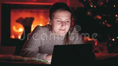 4k视频微笑的女孩在平板电脑上浏览互联网，同时躺在壁炉旁和发光的圣诞树