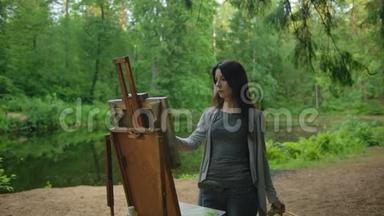 女艺术家穿着衬衫和牛仔裤在池塘附近的公园里画一幅<strong>风景画</strong>
