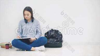 年轻的亚洲女孩坐在地板上，厌倦了写作，打哈欠。 <strong>所以</strong>，她拿着手机和耳机听着