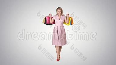 年轻的金发女孩穿着粉红色的连衣裙，展示给相机购物袋，并在渐变背景下行走。