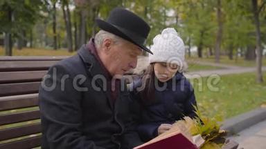 一个穿着经典服装的白种人成年男子和孙女坐在长凳上，读着一本红色封面的书