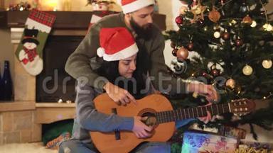 <strong>圣诞</strong>前<strong>夜圣诞</strong>老人戴`帽子的幸福夫妇的肖像。 男人教女人弹吉他。 <strong>圣诞</strong>树上的<strong>圣诞</strong>树