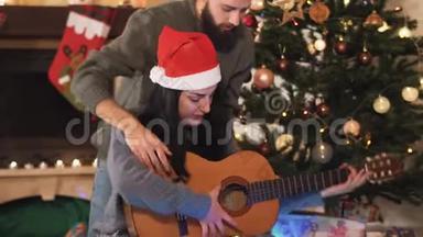 圣诞前夜圣诞老人戴`帽子的幸福夫妇的肖像。 男人教女人弹吉他。 圣诞树上的圣诞树