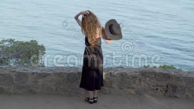 穿着黑色丝绸连衣裙和黑色帽子的长发女孩呆在蔚蓝海湾的小径上。 晴天，碧海松林..