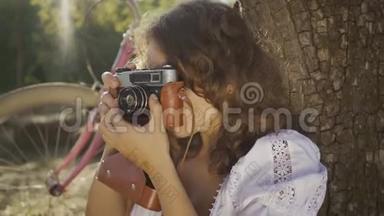 迷人的年轻摄影师，卷发<strong>坐在树下</strong>，在花园或公园里用旧相机拍照