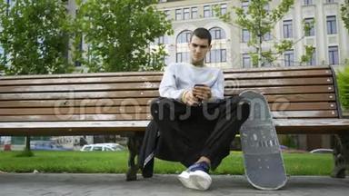 帅哥坐在公园的长凳上，用手机听音乐。 拐杖和滑板就在附近