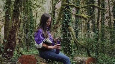 穿着<strong>运动夹克</strong>的女孩坐在森林里的树桩上，拧开热瓶的盖子从里面喝。