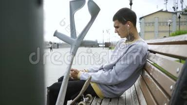 帅哥坐在公园的长凳上，用手机听音乐。 拐杖和滑板就在附近