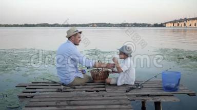 在河边野餐，爸爸和儿子钓完鱼后坐在河边的码头上，在芦苇丛中享受社交和度假的乐趣