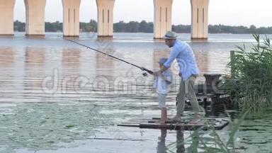 家庭日，渔夫父亲周末在河边的芦苇丛中教他的小儿子在码头上钓鱼