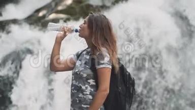 迷人的女孩喝着瓶子里的冷<strong>水</strong>，<strong>站</strong>在瀑布附近的石头上解渴