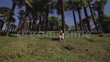穿着牛仔裤和夹克的漂亮女孩<strong>坐在</strong>棕榈<strong>树下</strong>的绿色草坪上