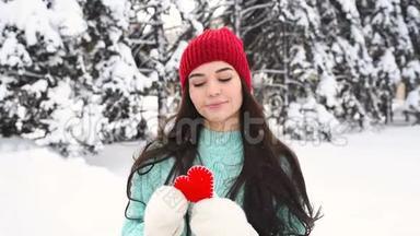 年轻漂亮的女孩穿着一件温<strong>暖</strong>的蓝色毛衣，戴着手套，背景是白雪覆盖的树木，抱着一颗<strong>红色</strong>的<strong>心</strong>，