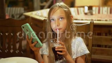十几岁的女孩喝着玻璃汽水，看着智能手机。 年轻女孩用手机喝汽水