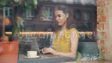 在咖啡馆里用笔记本电脑打字的漂亮女士微笑着在玻璃窗后面的桌子上