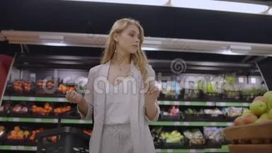 一个漂亮的女人走过超市，手里拿着一个篮子，看着水果的架子。 购买新鲜食品
