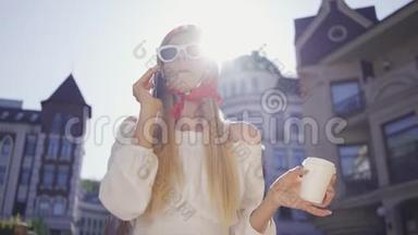 肖像时尚漂亮的年轻<strong>女人</strong>，带着一杯咖啡站在街上<strong>晒太阳</strong>，用手机说话
