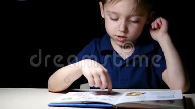 一个孩子晚上坐在一张灯点亮的桌子旁，读一本书，用手指指着书中的图画