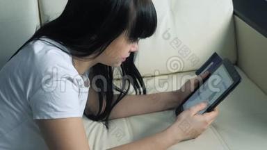女孩用数字平板电脑制作网上购物生活方式概念。 黑发女人躺在沙发上看网上商店