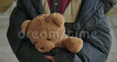 无家可归女孩的特写`她手里拿着泰迪熊。 疲惫的难民站在她可爱的玩具。