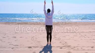 穿着帽衫和帽子的女人正在海沙滩做瑜伽，后景。