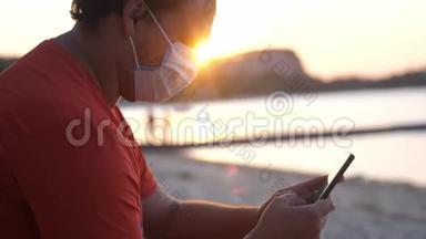 COVID-19. 男人戴着面具坐在海滩上，在日落时使用手机。 冠状病毒流行