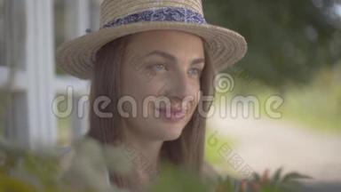 一个妩媚的年轻女子，戴着草帽，穿着白色连衣裙，坐在小村屋前。 农村