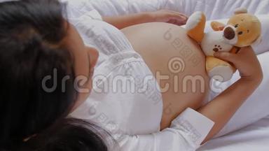 一位怀孕的拉丁美洲妇女坐在沙发上，抱着泰迪熊，把泰迪熊靠近她的腹部