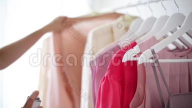 `白色衣架上穿粉色、米色和奶油色的女式服装。 特写镜头-一个女人拿起一件衬衫，溅起