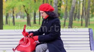 女人穿着红色的包在秋天公园的长凳上看东西。 教师节