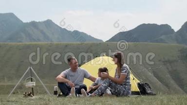 一个幸福的家庭和一个孩子在山上的帐篷前休息，喝着热水瓶里的茶