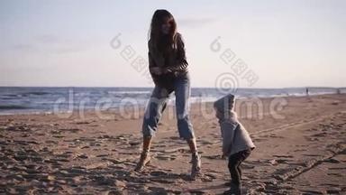 有趣的妈妈和穿着暖和的衣服的小宝宝在海边的沙滩上跳舞，跳着跳着玩