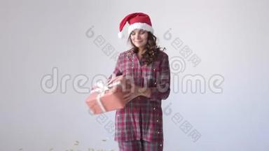 穿着圣诞帽和睡衣的年轻女子在白色背景上带着礼品盒跳舞。 女人手里的小礼物