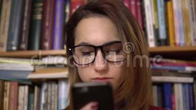 一个年轻的女人正在图书馆看智能手机。 戴眼镜的女人仔细地看着手机特写。 在里面