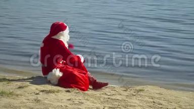 圣诞老人坐在河边或湖边，拿着一袋礼物。 圣诞老人看着远处的水。 圣诞老人