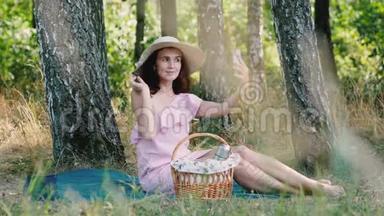 野餐时间。 年轻漂亮的女孩，戴着圆帽子，坐在格子上，摆出自拍的姿势。 公园的户外活动