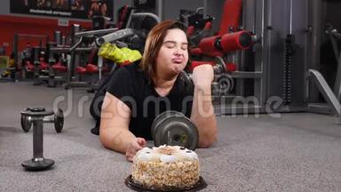 运动与不健康的食物。 年轻的胖女人把杠铃放下，拿一个大蛋糕。 年轻女子