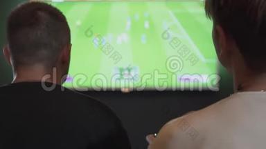 后景两人在游戏室玩电子游戏坐在沙发上。 踢足球或踢足球的朋友