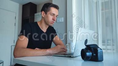 年轻的摄影师使用笔记本电脑处理图像在他的笔记本电脑和喝咖啡在家里。 外部硬盘摄像头