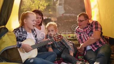 旅游家庭在森林徒步旅行的露营帐篷里用吉他演<strong>唱歌</strong>曲。 友好的家庭在露营时<strong>唱歌</strong>