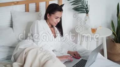 穿着浴袍的年轻女子躺在床上，用笔记本电脑在网上工作或学习