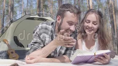 肖像胡子男和可爱的年轻女子躺在森林里的帐篷里看书。 爱