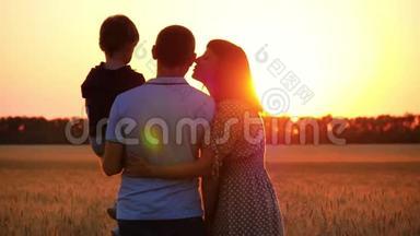 幸福的一家人看着夕阳，站在麦田上。 抱着孩子的男人.. 女人拥抱男人，亲吻他。