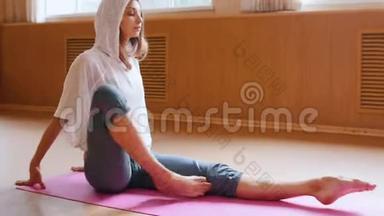 穿着白色连帽衫、坐在瑜伽垫上进行腿部练习的年轻苗条女子<strong>舞蹈工作室</strong>