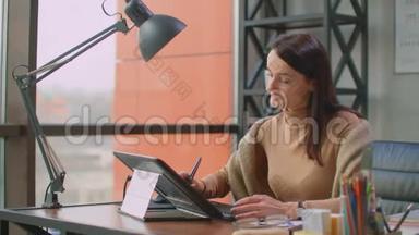 <strong>办公室里</strong>的一位专业设计师在<strong>办公室里</strong>坐着一个巨大的窗户，在平板电脑上用手写笔画画