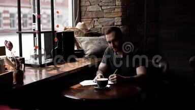 在咖啡馆里，一个穿着纹身手臂坐在窗前的粗鲁男人在垫子上做笔记