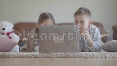 肖像<strong>可爱</strong>的<strong>双胞胎</strong>男孩和女孩躺在地毯上，看着电脑上有趣的东西，笑着，