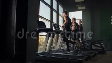 两个男人在健身房一个大窗户的背景下在跑步机上跑步。 参加健身运动的年轻人