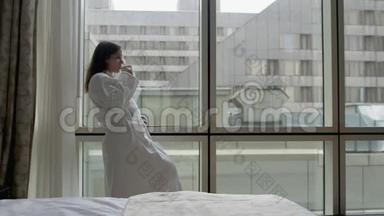穿着<strong>白色浴袍</strong>的年轻女子站在酒店公寓的窗户旁，手里拿着智能手机和一杯咖啡