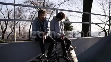 两个朋友滑板车和bmx骑手正坐在城市滑板公园的高栏杆上，使用他们的滑板车